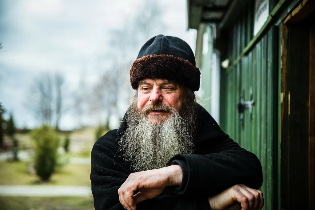 Starszy mężczyzna z długą siwą brodą i w futrzanej czapce opiera się o drewnianą poręcz, uśmiechając się lekko, w tle zielony barak oraz rozmyte drzewa i trawnik.