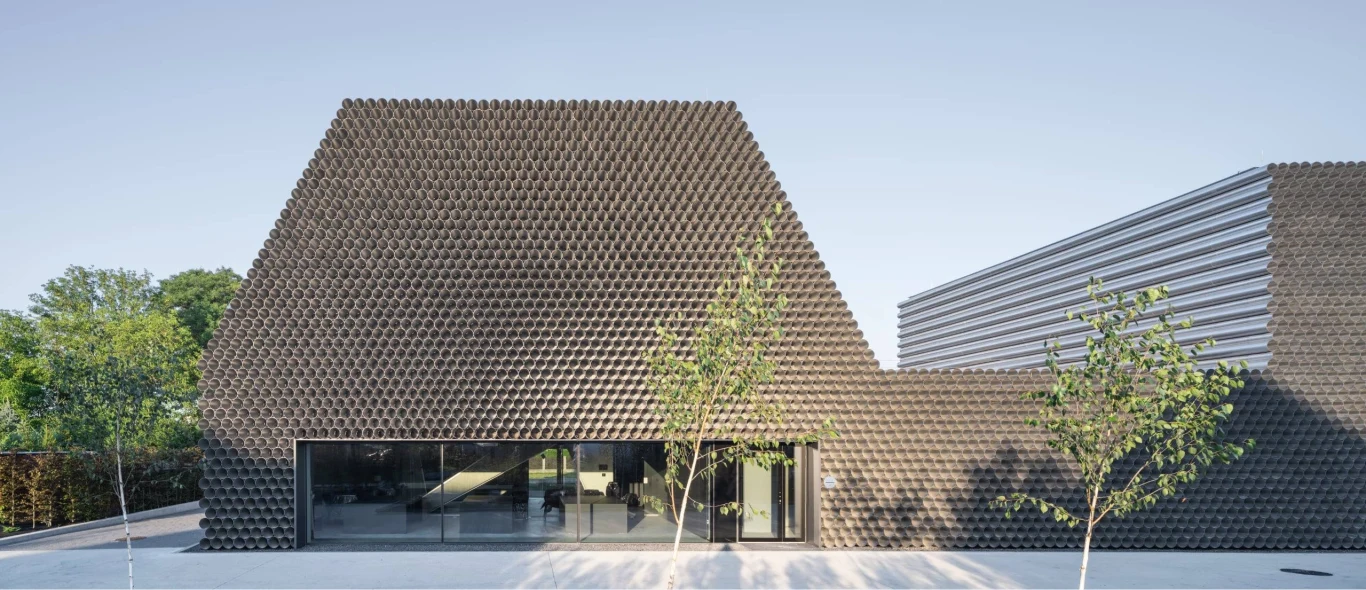 Nowoczesny budynek o oryginalnej bryle, z elewacją pokrytą ciemnymi, geometrycznymi elementami oraz dużym przeszkleniem na parterze; przed budynkiem rosną młode drzewa, a całość otacza czysta przestrzeń.