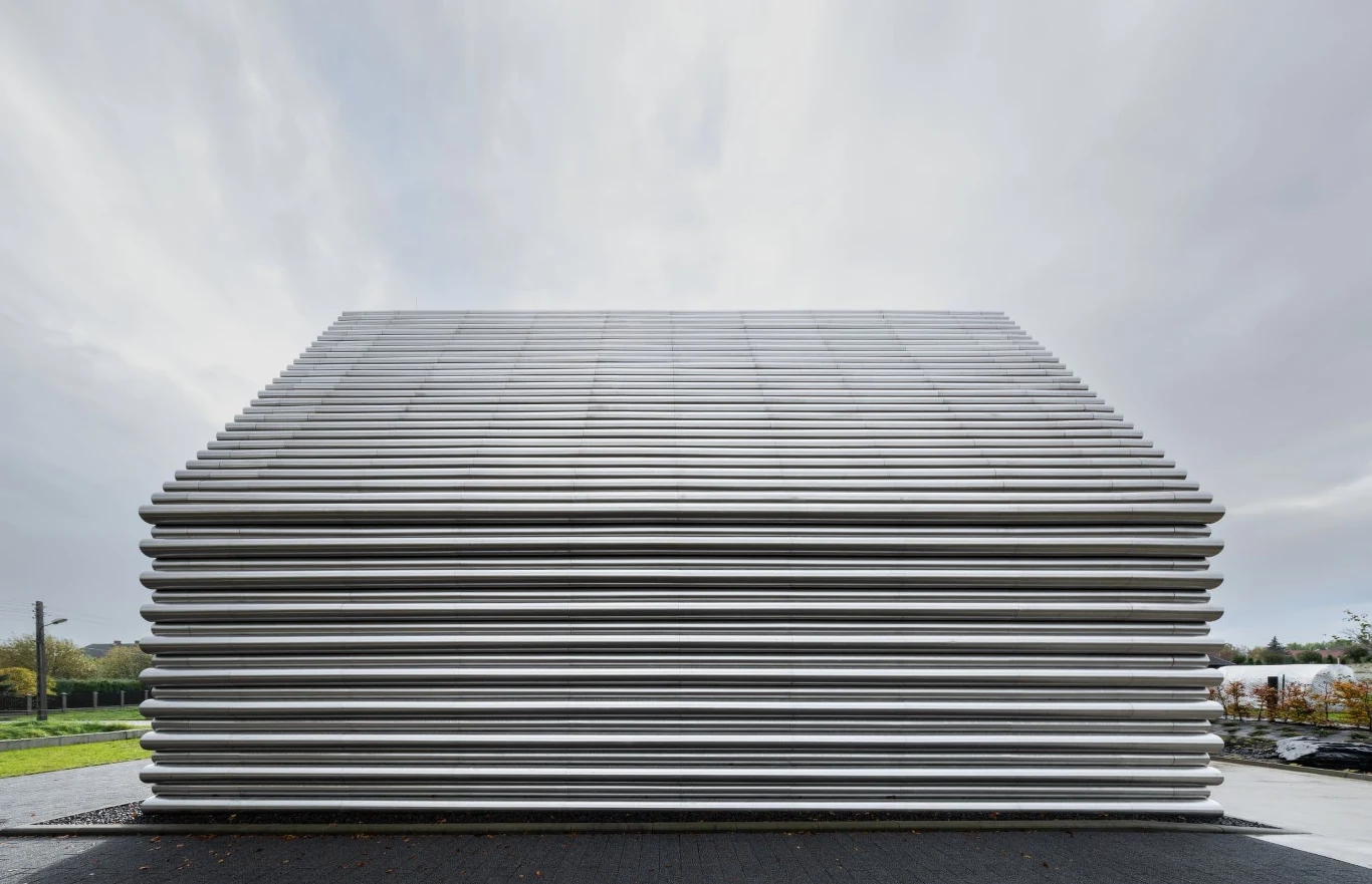 Nowoczesny budynek o industrialnym wyglądzie, którego elewację tworzą poziome, równoległe aluminiowe listwy, otoczony niewielką ilością zieleni i z pochmurnym niebem w tle.