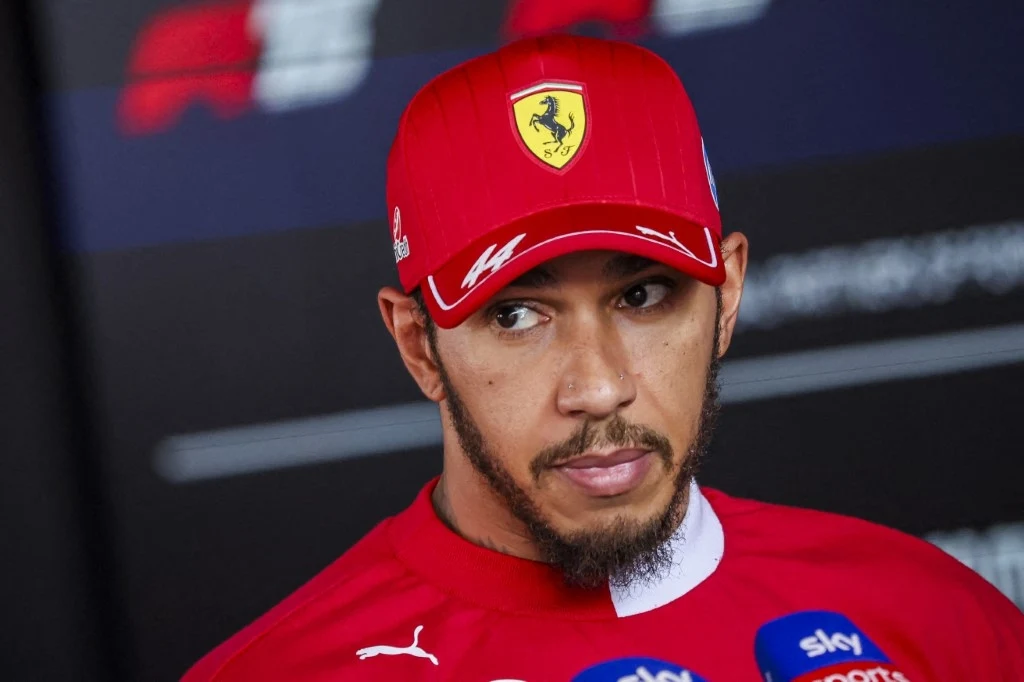 Mężczyzna w czerwonej czapce i koszulce zespołu Ferrari odpowiada na pytania podczas konferencji prasowej, przed nim mikrofony z logo różnych mediów sportowych.