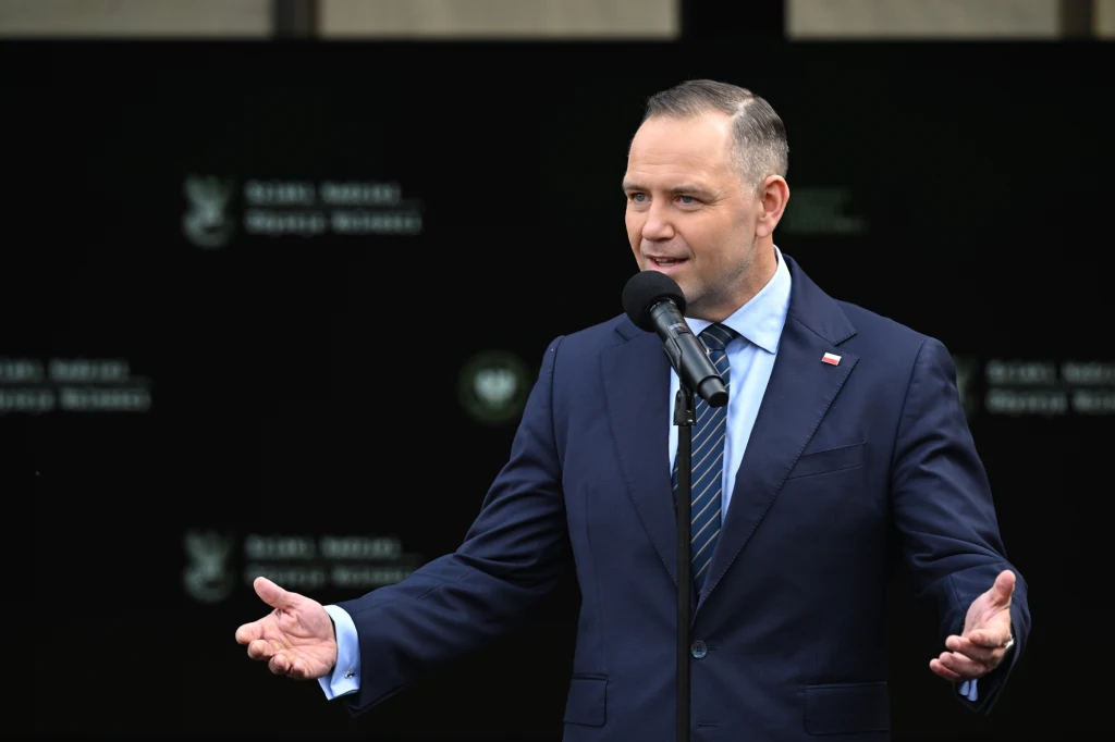 Karol Nawrocki Mężczyzna w garniturze wygłasza przemówienie na zewnątrz, stojąc za mównicą z mikrofonem, gestykulując dłońmi, w tle ciemne tło z niewyraźnym napisem.