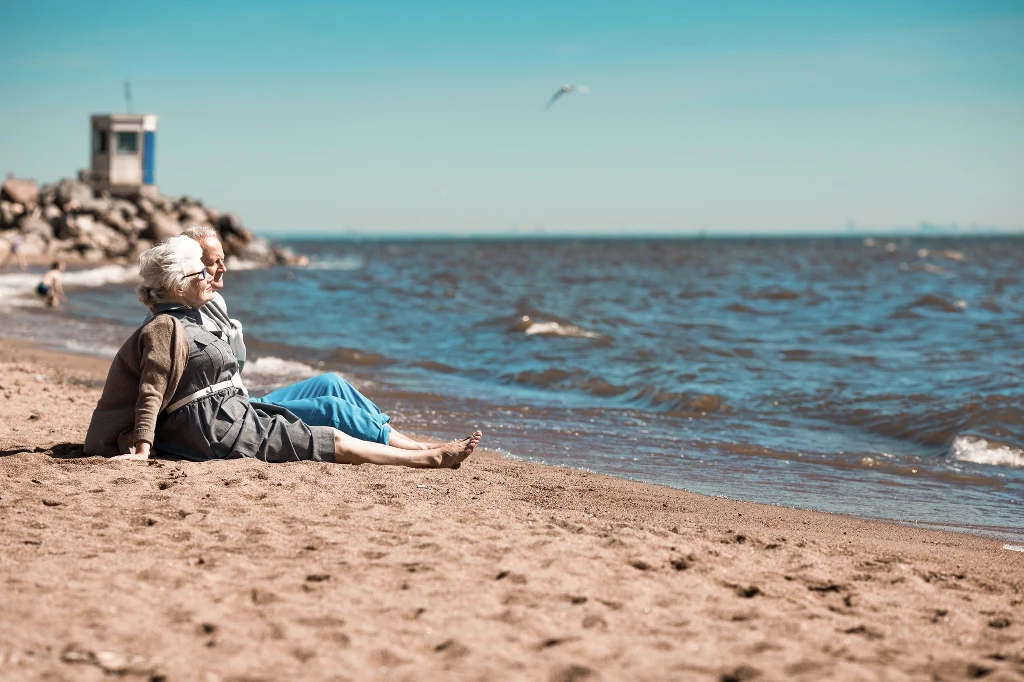Dwie starsze osoby siedzące na piaszczystej plaży, zwrócone w stronę morza, cieszące się spokojnym, słonecznym dniem; w tle widoczne fale oraz niewielki budynek przy kamienistym nabrzeżu.