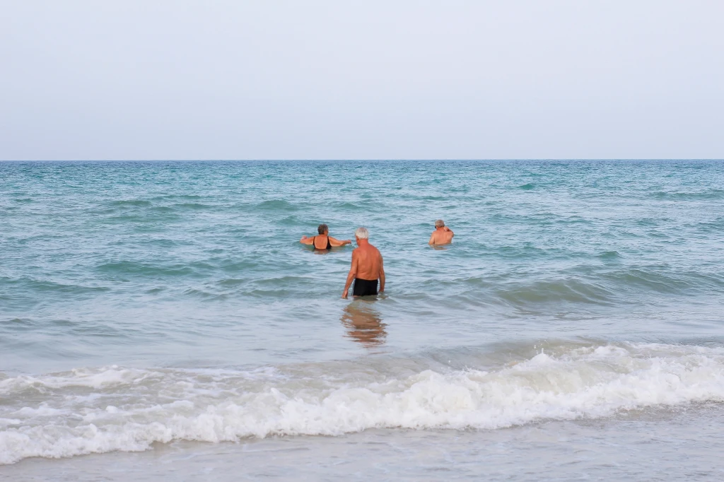 Trzy osoby kąpiące się w spokojnym morzu, nieco oddalone od brzegu, na pierwszym planie lekkie fale docierające do piaszczystej plaży, spokojna atmosfera letniego wypoczynku.