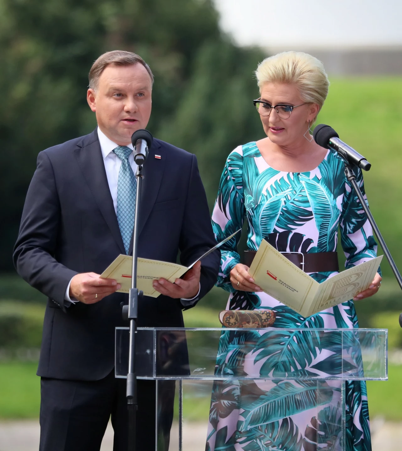 Agata Duda jest fanką kwiecistych sukienek Dwoje dobrze ubranych dorosłych stoi przy mównicy z mikrofonami, trzymając otwarte dokumenty, na tle zieleni i drzew, skupieni na czytaniu lub przemówieniu.
