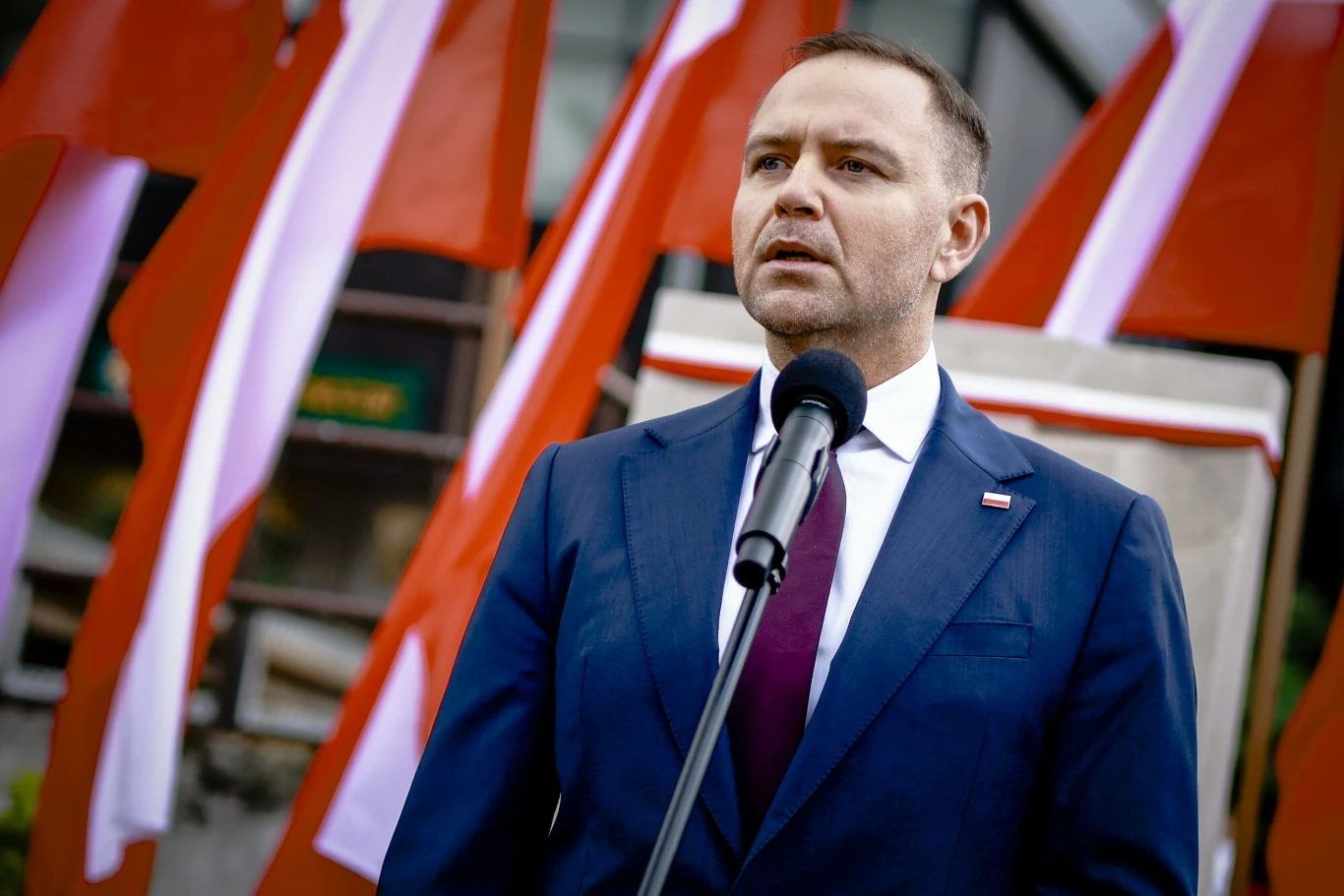Mężczyzna w garniturze stojący przed mikrofonem, w tle kilka polskich flag.