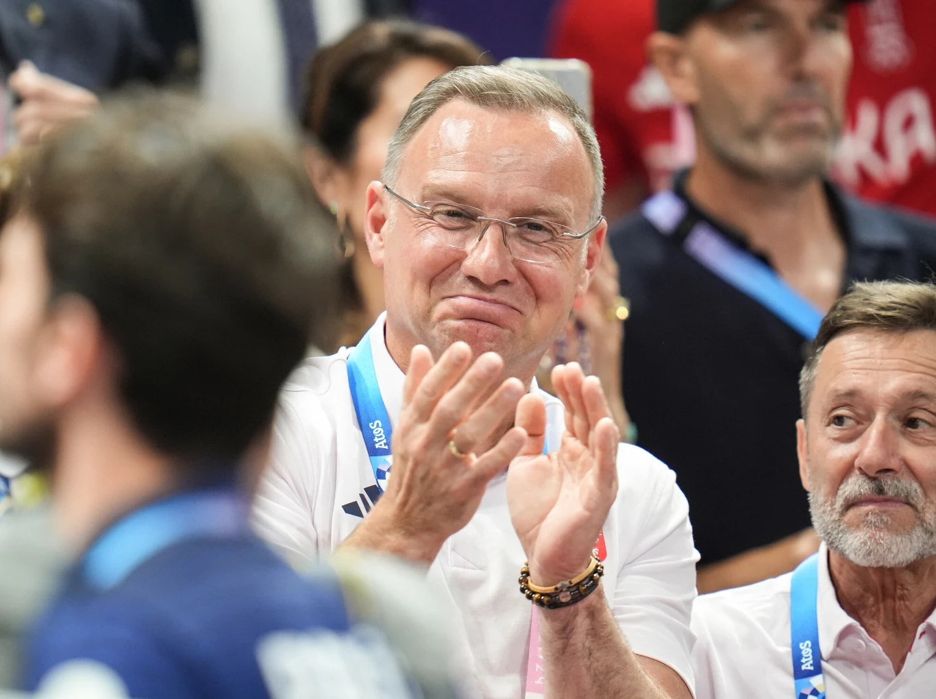 Mężczyzna w okularach z uśmiechem bije brawo, wokół niego stoją inni ludzie, niektórzy mają identyfikatory na szyjach, atmosfera zasugerowana jest jako pozytywna i oficjalna.