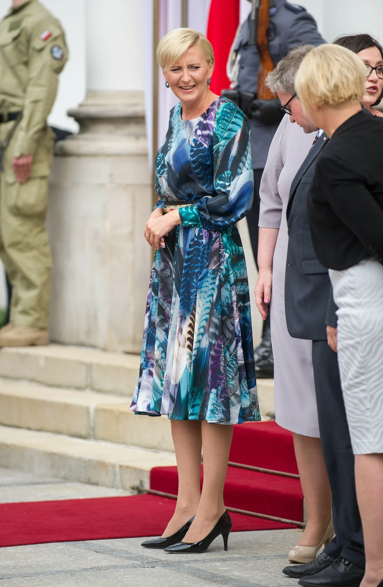 Agata Duda w 2016 roku Elegancko ubrana kobieta w kolorowej sukience stoi na schodach przed wejściem do budynku, w towarzystwie innych osób, na tle czerwonego dywanu; otoczenie sugeruje oficjalne wydarzenie.