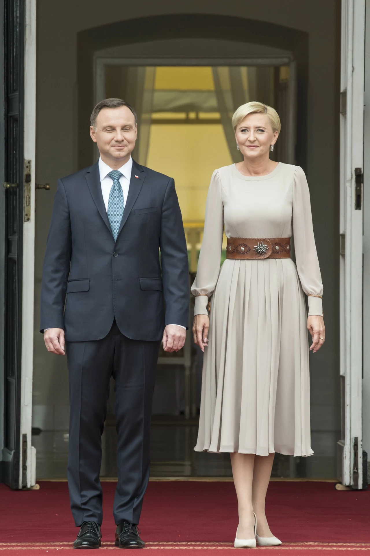 Para prezydencka witająca księcia Williama i księżną Kate Elegancko ubrany mężczyzna i kobieta stoją razem przed wejściem do budynku na czerwonym dywanie, oboje uśmiechnięci, mężczyzna w garniturze z krawatem, kobieta w jasnej sukience z szerokim paskiem.