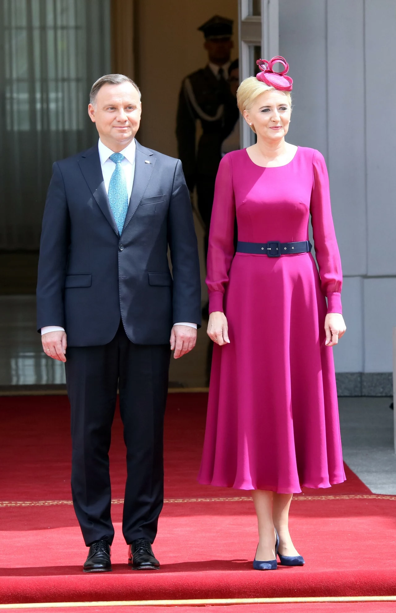 Para prezydencka w 2019 roku Elegancko ubrany mężczyzna w garniturze i kobieta w fioletowej sukni z ozdobnym nakryciem głowy stoją razem na czerwonym dywanie przed wejściem do budynku, w tle widoczny strażnik.
