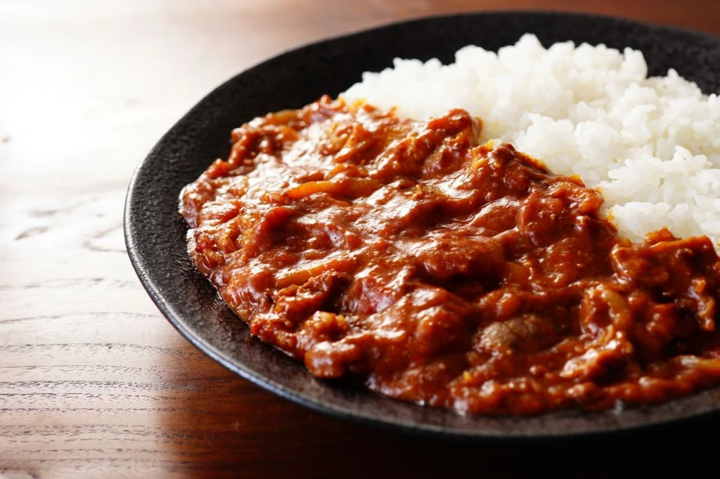Porcja białego ryżu ułożona obok ciemnego, gęstego curry o czerwono-brązowej barwie, całość podana na czarnym talerzu umieszczonym na drewnianym stole.