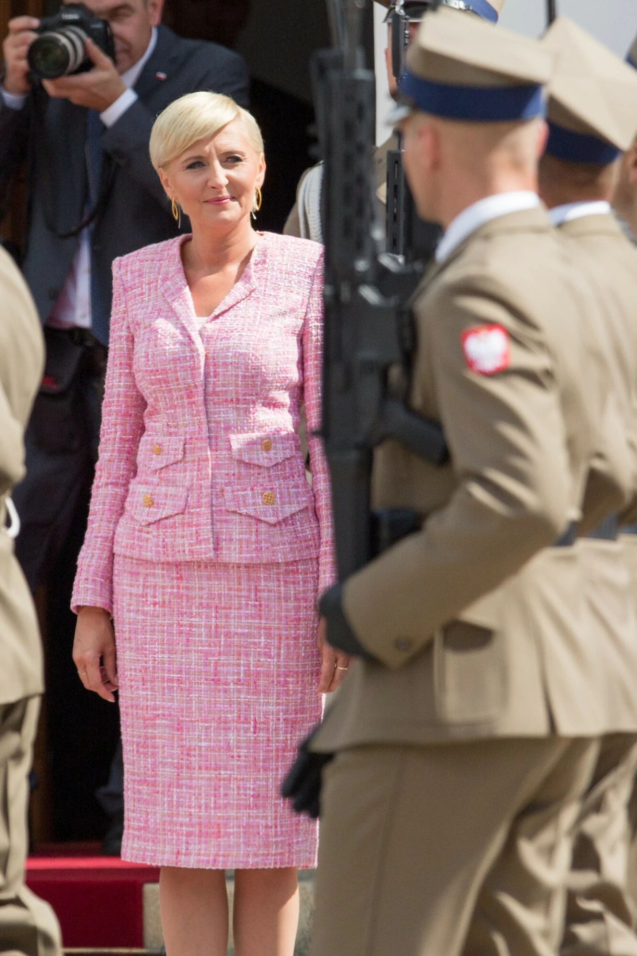 Agata Duda w tweedowej garsonce Kobieta w eleganckim różowym kostiumie stoi zwrócona w stronę żołnierzy w mundurach, z których jeden trzyma broń. W tle widać fotografa z aparatem fotograficznym.