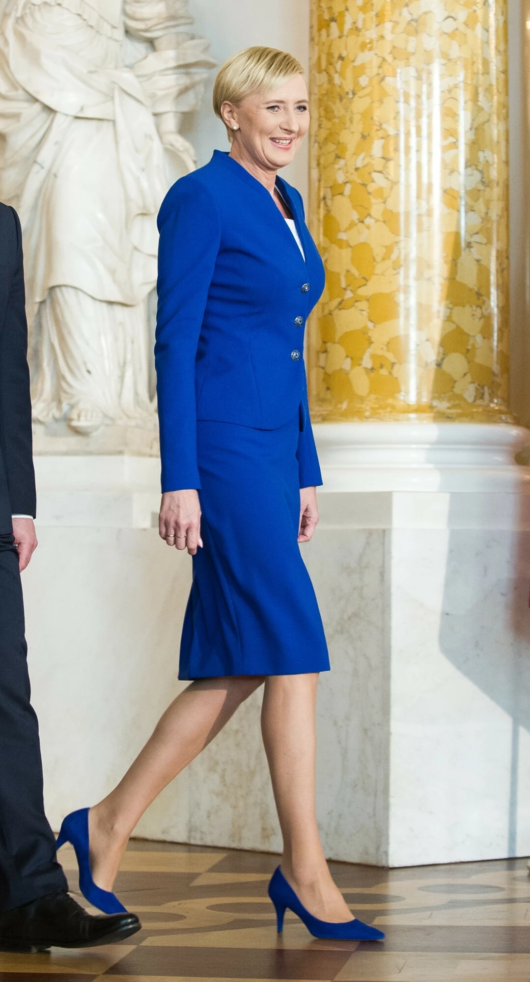 Agata Duda w 2017 roku Elegancko ubrana kobieta w niebieskim garniturze i szpilkach zmierza przez bogato zdobione wnętrze z marmurową kolumną i klasycznymi rzeźbami w tle, uśmiecha się, sprawia wrażenie pewnej siebie i zadowolonej.