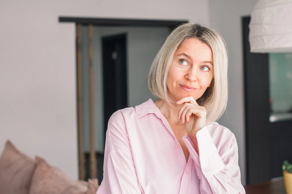Kobieta z blond włosami ubrana w jasnoróżową koszulę stoi w jasnym wnętrzu, delikatnie uśmiechnięta, z ręką opartą pod brodą, sprawia wrażenie zamyślonej.