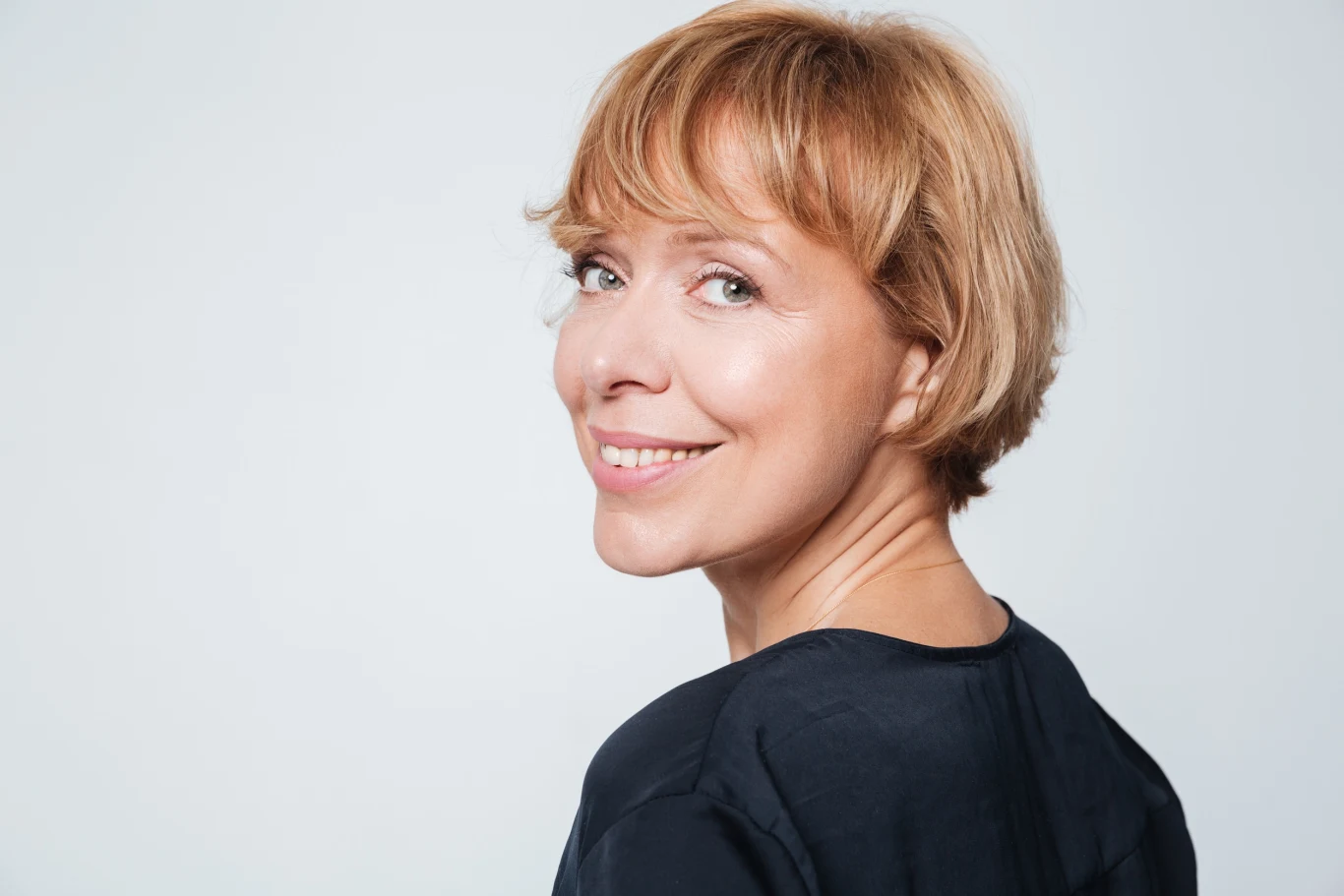 Grzywka to element fryzury, który może całkowicie odmienić jej charakter Uśmiechnięta kobieta o krótkich, jasnobrązowych włosach, ubrana w ciemną bluzkę, patrzy przez ramię na wprost obiektywu, na jasnym tle.