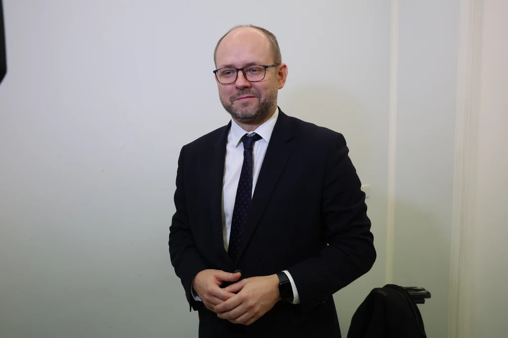 Karol Nawrocki powołał Marcina Przydacza na ministra odpowiedzialnego za sprawy zagraniczne i Polonię Mężczyzna w okularach ubrany w ciemny garnitur, białą koszulę oraz krawat stoi na tle jasnej ściany, uśmiecha się delikatnie i patrzy do obiektywu.
