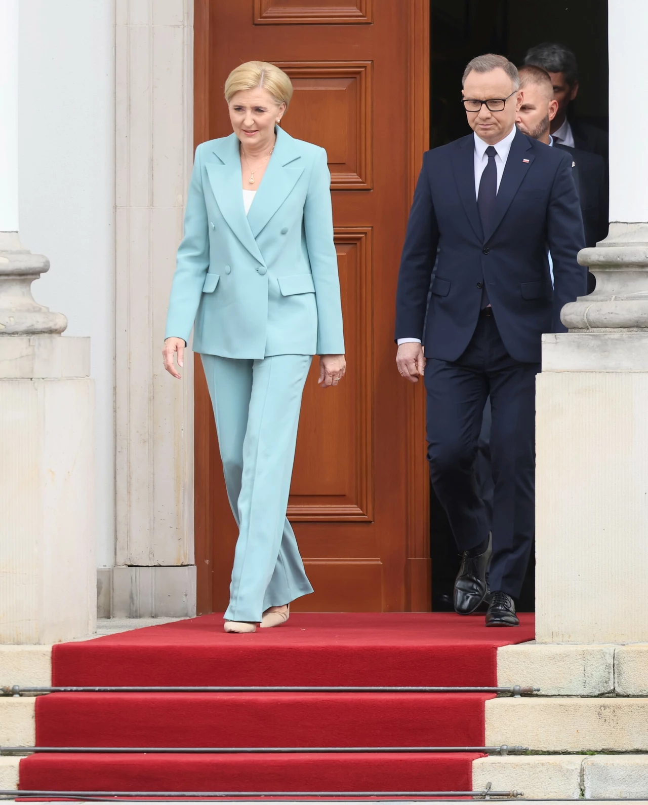 Agata Duda w czerwcu 2025 roku Kobieta w jasnoniebieskim garniturze oraz mężczyzna w granatowym garniturze schodzą po czerwonym dywanie przed budynkiem z drewnianymi drzwiami.