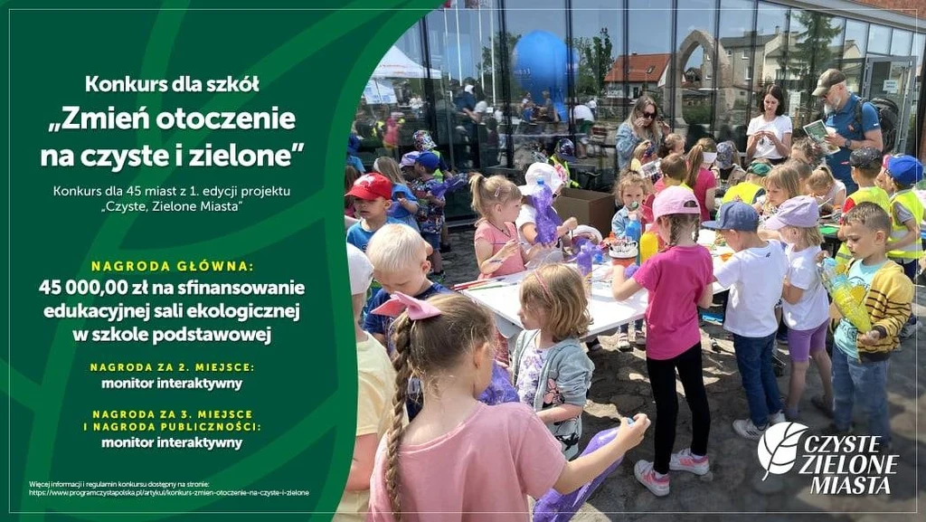 Konkurs dla szkół "Zmień otoczenie na czyste i zielone" Grupa dzieci skupiona przy stole plastycznym podczas zajęć edukacyjnych na świeżym powietrzu, wokół stoi kadra opiekuńcza i rodzice, po lewej stronie zielony plakat konkursowy z informacjami o konkursie "Czyste Zielone Miasta" oraz nagrodach.