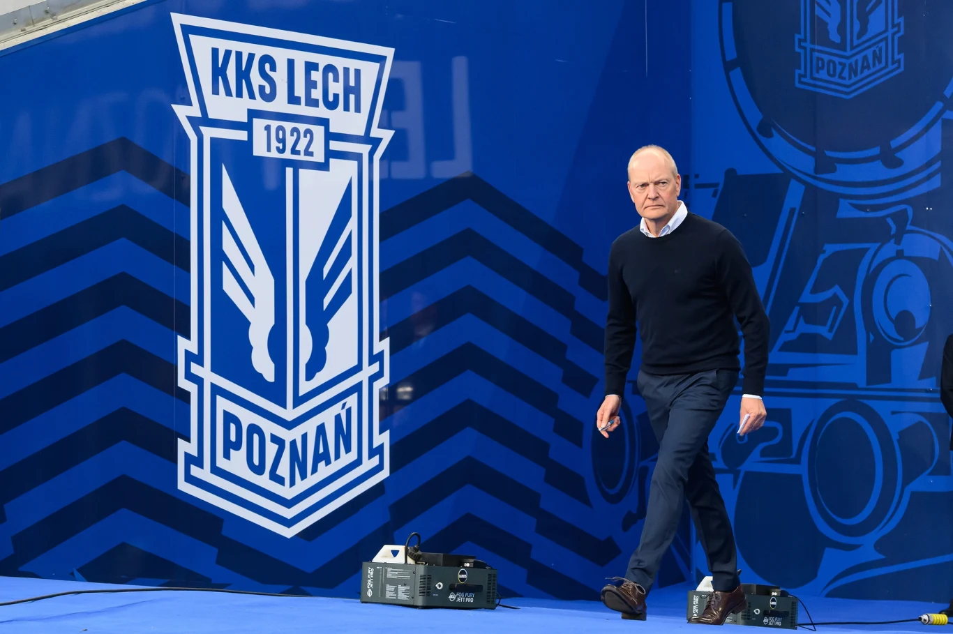 Niels Frederiksen, trener Lecha Poznań Mężczyzna w ciemnym swetrze i spodniach idzie obok dużego niebieskiego emblematu klubu piłkarskiego KKS Lech Poznań, na tle ściany ozdobionej klubowymi elementami graficznymi.