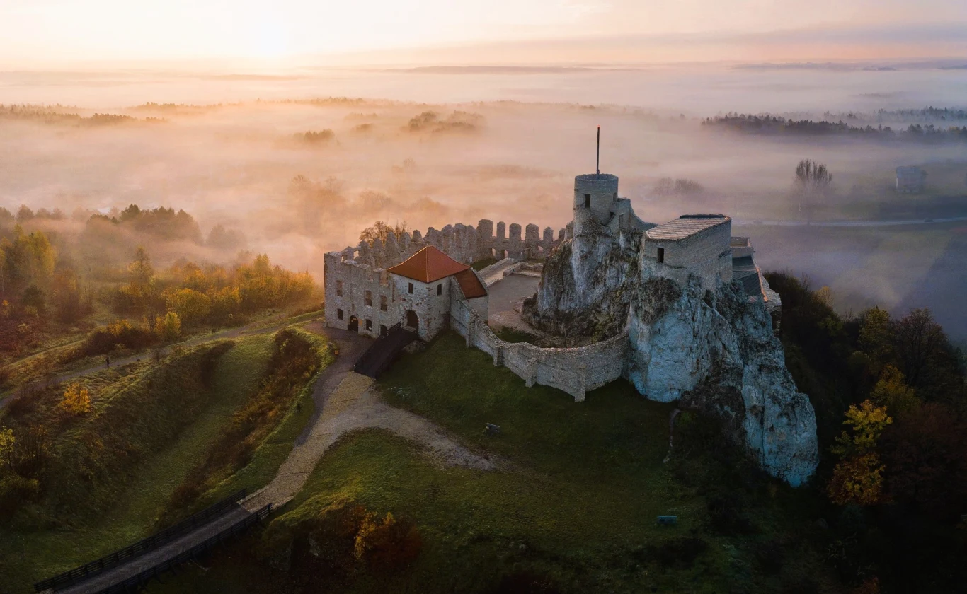Zamek Rabsztyn już teraz stanowi jedną z pereł Szlaku Orlich Gniazd, a wkrótce stanie się jeszcze ciekawszy. Podano szczegóły na temat inwestycji. Zamek na wzgórzu otoczony poranną mgłą, z wyraźnie widocznymi murami obronnymi i kamienną basztą, otoczony jesienną roślinnością, z panoramą rozciągającą się w tle.