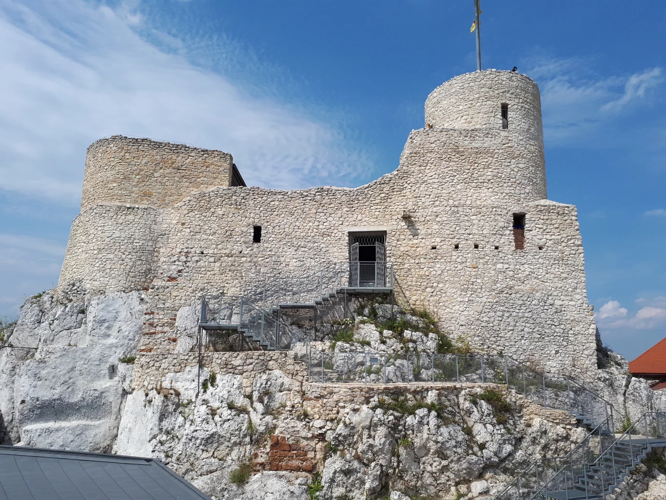 Kamienny zamek na skale z dobrze zachowaną cylindryczną wieżą, białe mury oraz wejście dostępne po metalowych schodach, błękitne niebo w tle.