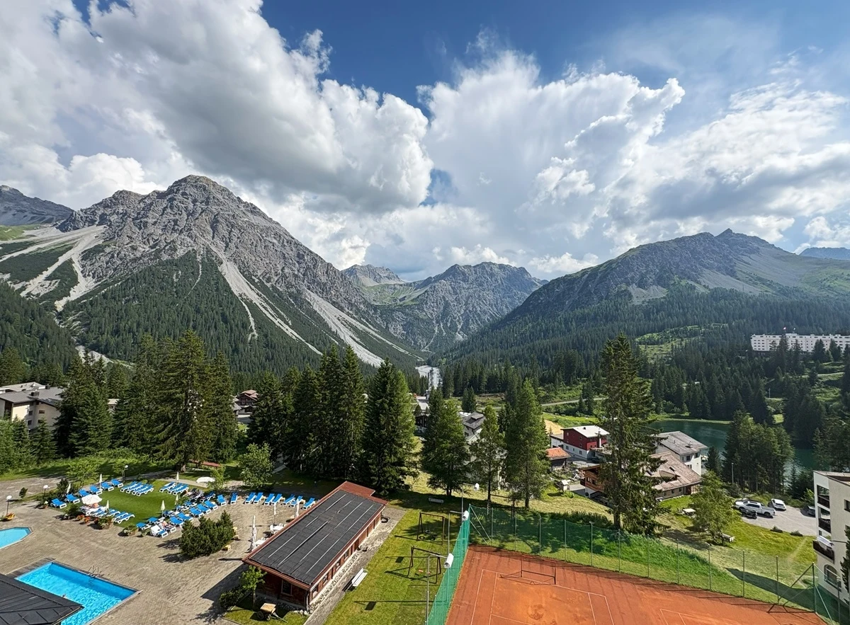 Szwajcaria nie tylko dla amatorów białego szaleństwa. Kraj zachwyca również latem Górska panorama z wyraźnie zarysowanymi szczytami, gęstym lasem iglastym i rozproszoną zabudową wypoczynkową, na pierwszym planie widoczne są baseny, kort tenisowy oraz budynki rekreacyjne