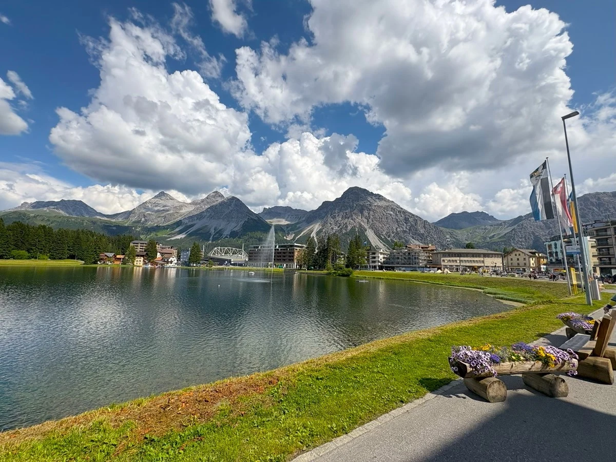 Arosa: jeden z najpopularniejszych alpejskich kurortów Górskie jezioro otoczone zielenią, w tle wysokie, majestatyczne szczyty oraz zabudowania na brzegu, po prawej stronie flagi i ławki z kwiatami pod błękitnym niebem z dużymi chmurami.