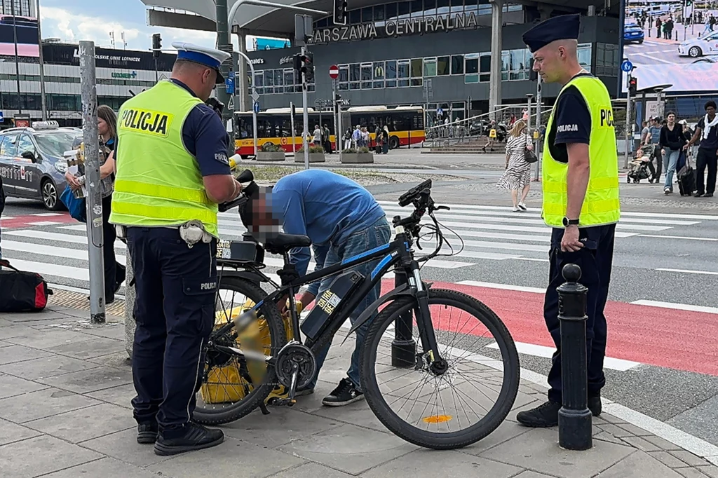 Dwóch policjantów w żółtych kamizelkach kontroluje mężczyznę przy rowerze, stojącego na chodniku obok ulicy, w tle widać budynek dworca Warszawa Centralna oraz przejście dla pieszych i przejeżdżające samochody.