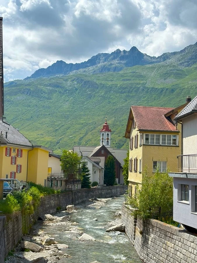 Spacer po Andermatt, położonym w pobliżu Przełęczy Świętego Gotarda Górska rzeka płynąca przez miasteczko otoczona kolorowymi budynkami, w tle góry porośnięte zielenią i niebo z chmurami, pośrodku widoczna wieża kościoła z czerwoną kopułą.