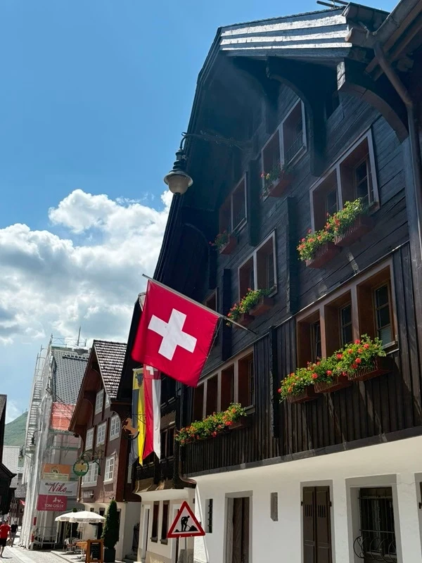Andermatt leży pośrodku alpejskich przełęczy Drewniany budynek z licznymi balkonami ozdobionymi skrzynkami pełnymi kwiatów, na głównym balkonie wywieszona flaga Szwajcarii, ka ulicy widoczna flaga Niemiec i znak drogowy ostrzegający przed dziećmi, w tle więcej tradycyjnych zabudowań oraz niebo z ...