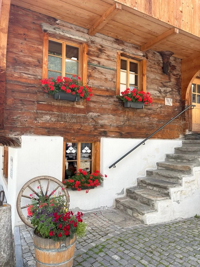 Charakterystyczna architektura, góry i bliskość natury? Tak, to Szwajcaria Drewniana elewacja domu ozdobiona skrzynkami z czerwonymi kwiatami na parapetach okien, kamienne schody prowadzące na piętro, beczka z kwiatami oraz stara drewniana część koła ustawiona przy elewacji.