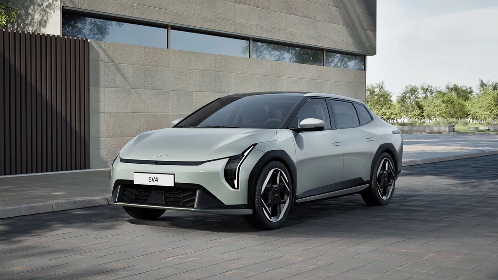 Kształty modelu EV4 zostały zaprojektowane tak, aby uzyskać jak najniższy współczynnik oporu powietrza. srebrny samochód elektryczny o nowoczesnym, dynamicznym designie zaparkowany przed budynkiem z dużymi oknami, w tle widoczne drzewa i minimalistyczna architektura