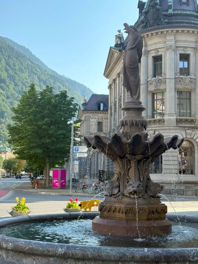 Kamienna fontanna z elegancką rzeźbą kobiety na szczycie, ozdobiona misami z tryskającą wodą na tle klasycznego budynku z bogatą architekturą i górskim krajobrazem w tle.