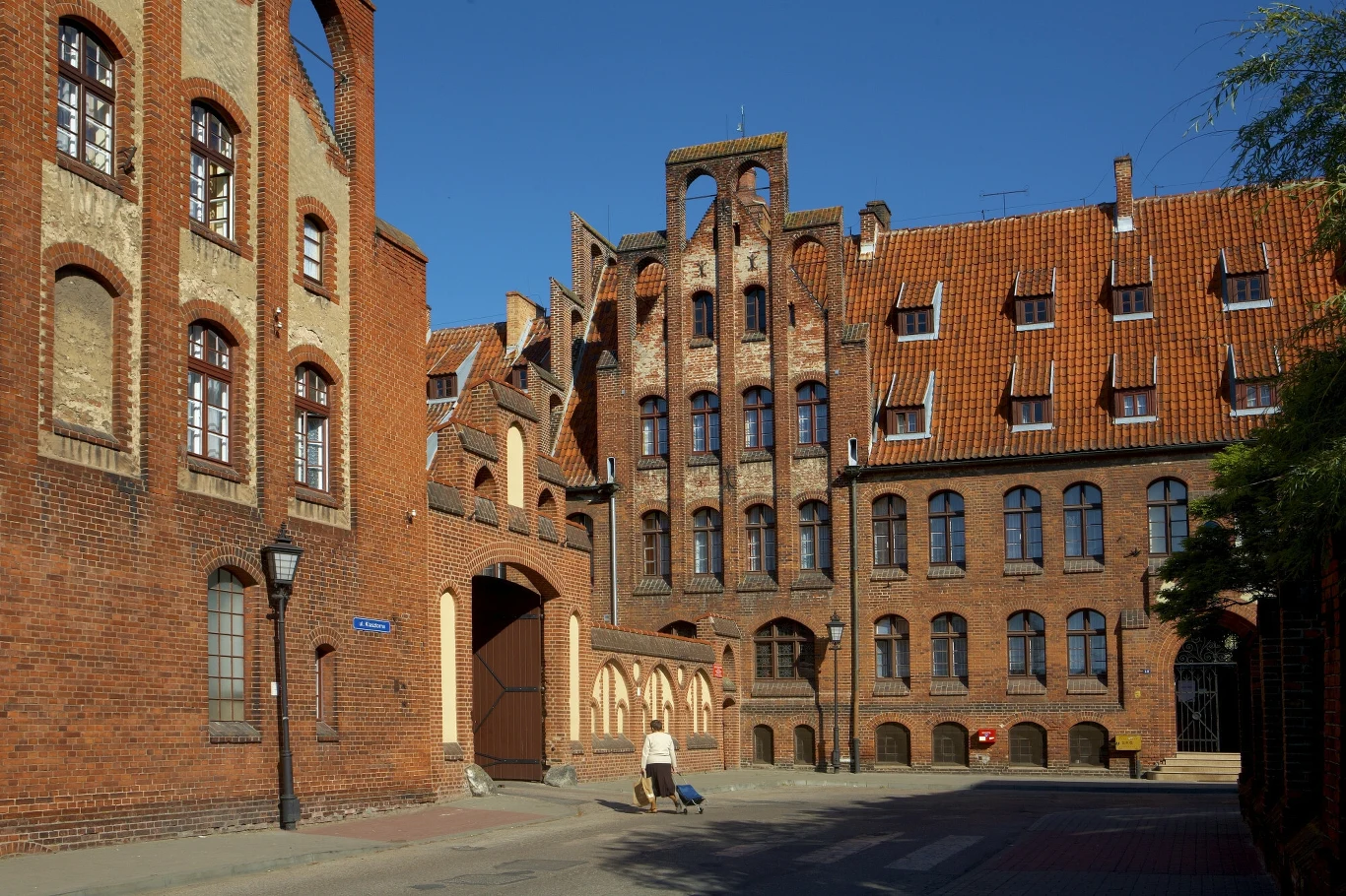 Ceglany kompleks budynków o historycznej architekturze z ozdobnymi detalami i stromymi dachami, na tle bezchmurnego nieba, ulicą przechodzi pieszy z walizką na kółkach.