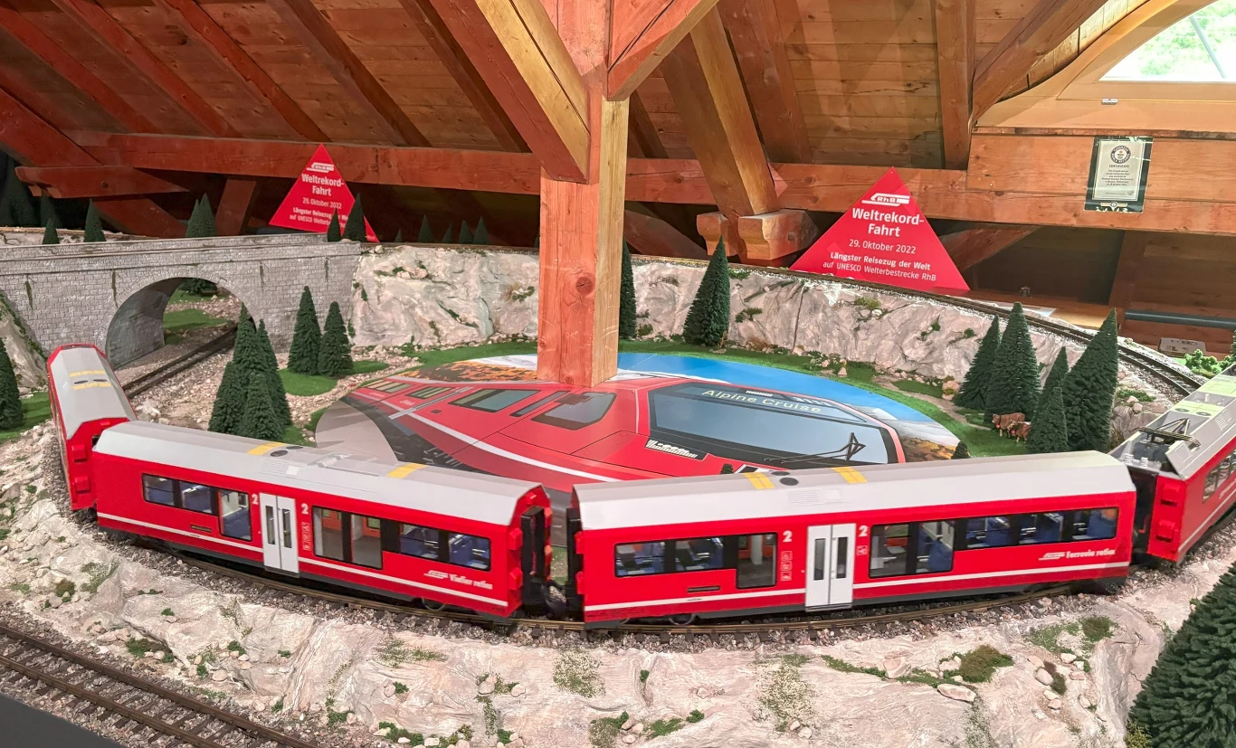 Bergün. Z wizytą w Muzeum Kolei Albula Czerwony model pociągu jadący po makiecie kolejowej otoczonej miniaturowymi drzewami i tunelami skalnymi, w tle drewniane bele poddasza.