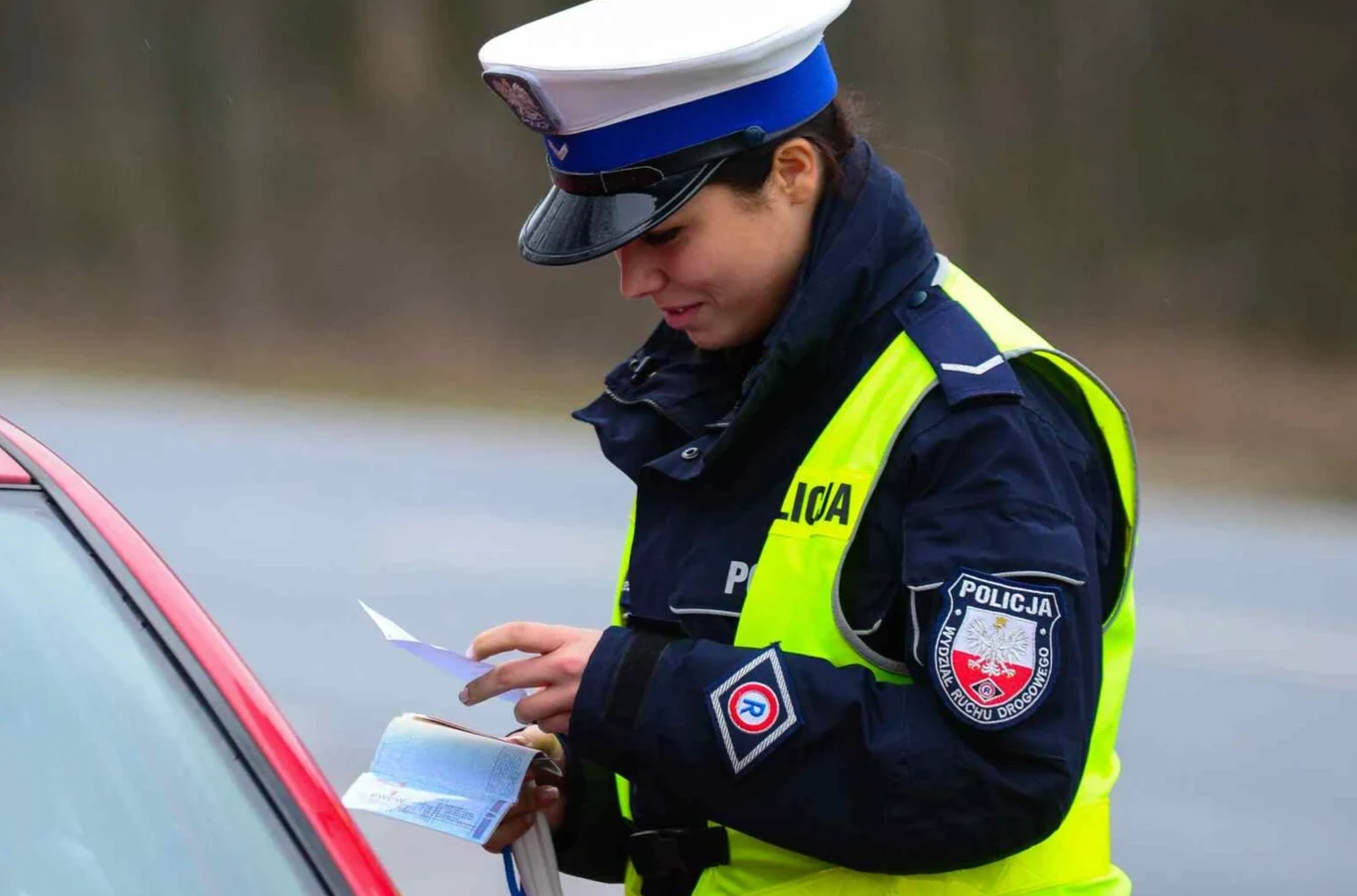 Co oznacza pole S.1 w dowodzie rejestracyjnym? policjantka w mundurze i czapce policyjnej sprawdza dokumenty kierowcy przy czerwonym samochodzie, trzymając papiery i notes na tle rozmytego, szarego otoczenia