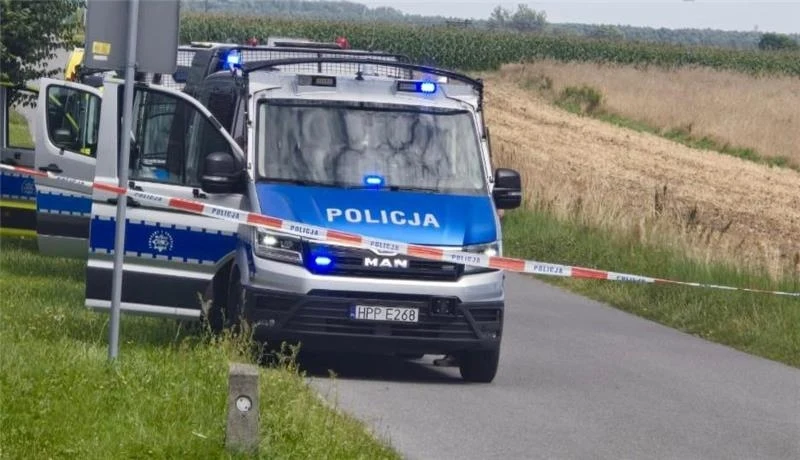 Radiowóz policyjny z włączonymi sygnałami świetlnymi stoi na wiejskiej drodze, odgrodzony taśmą policyjną, z polami i fragmentem pojazdu policyjnego w tle.
