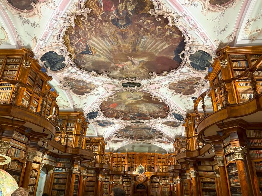 Barokowa biblioteka z bogato zdobionym sufitem ozdobionym freskami, wnętrze wypełnione drewnianymi regałami pełnymi książek, licznymi balkonami i galeriami.