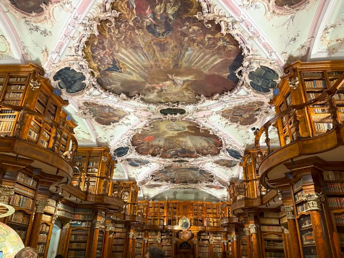 Wnętrze biblioteki opactwa św. Galla w Sankt Gallen Barokowa biblioteka z bogato zdobionym sufitem ozdobionym freskami, wnętrze wypełnione drewnianymi regałami pełnymi książek, licznymi balkonami i galeriami.