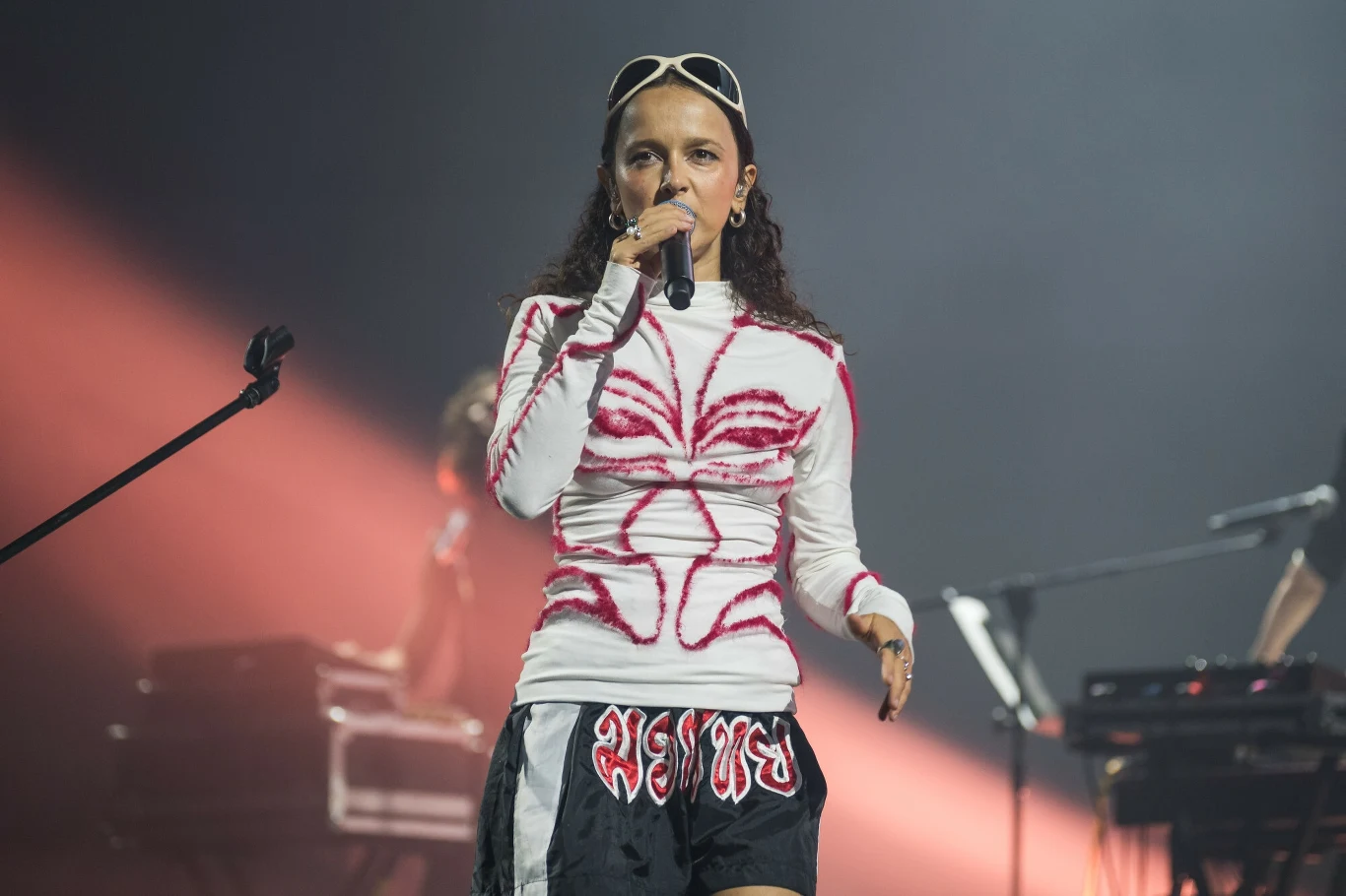 Monika brodka podczas koncertu Męskiego Grania we Wrocławiu Wokalistka występująca na scenie z mikrofonem, ubrana w biały długi rękaw z różowym wzorem i czarne szorty, w tle widoczna scenografia koncertowa oraz światła sceniczne.