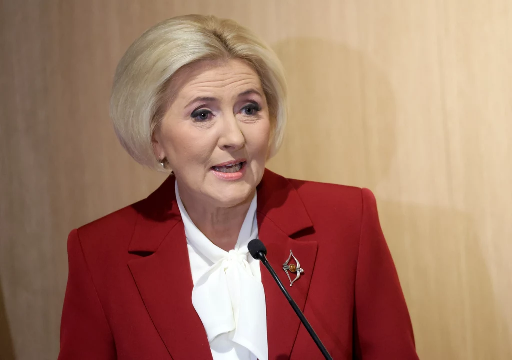 Agata Duda podczas otwarcia najnowocześniejszej w Polsce Poradni Chorób Piersi w Wojskowym Instytucie Medycznym Państwowym Instytucie Badawczym Kobieta w średnim wieku o jasnych włosach w eleganckim czerwonym żakiecie i białej bluzce stoi przy mównicy z mikrofonem, przemawia w jasnym, formalnym wnętrzu.