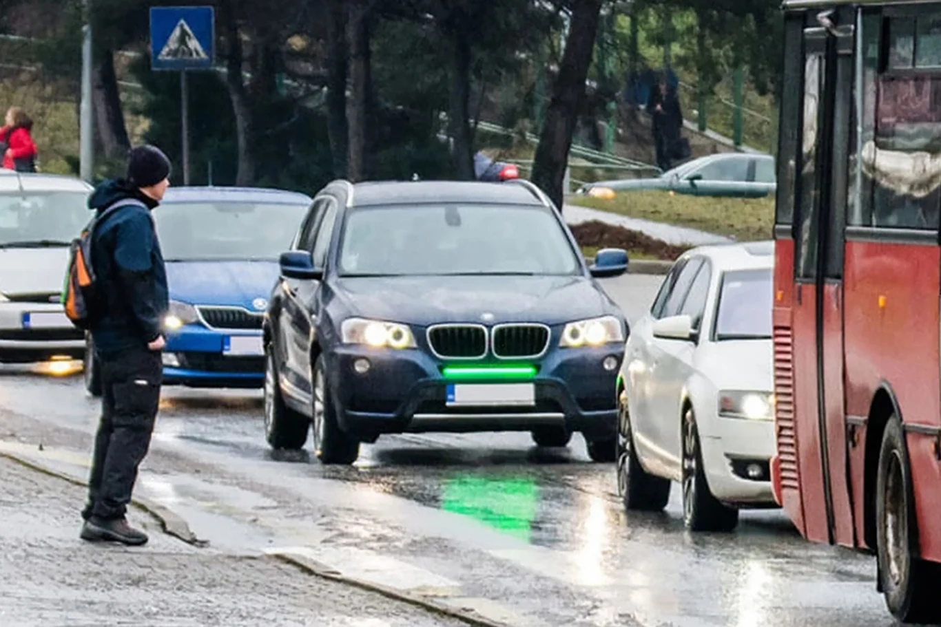 Przednie światła stopu pomogą zapobiegać kolizjom? | Fot. frontbrakelights.com Ruchliwe skrzyżowanie, samochód BMW z jasnozielonym światłem na przednim zderzaku, stojący przed przejściem dla pieszych na mokrej jezdni, obok autobusu i przechodnia czekającego na możliwość przejścia.