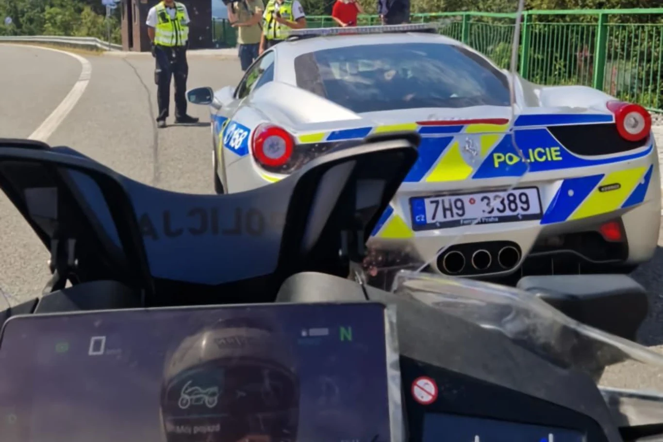 Radiowóz policyjny Ferrari zatrzymany na poboczu, otoczony przez kilku funkcjonariuszy w kamizelkach odblaskowych, w przednim planie widoczna jest kierownica innego pojazdu z cyfrowym wyświetlaczem.