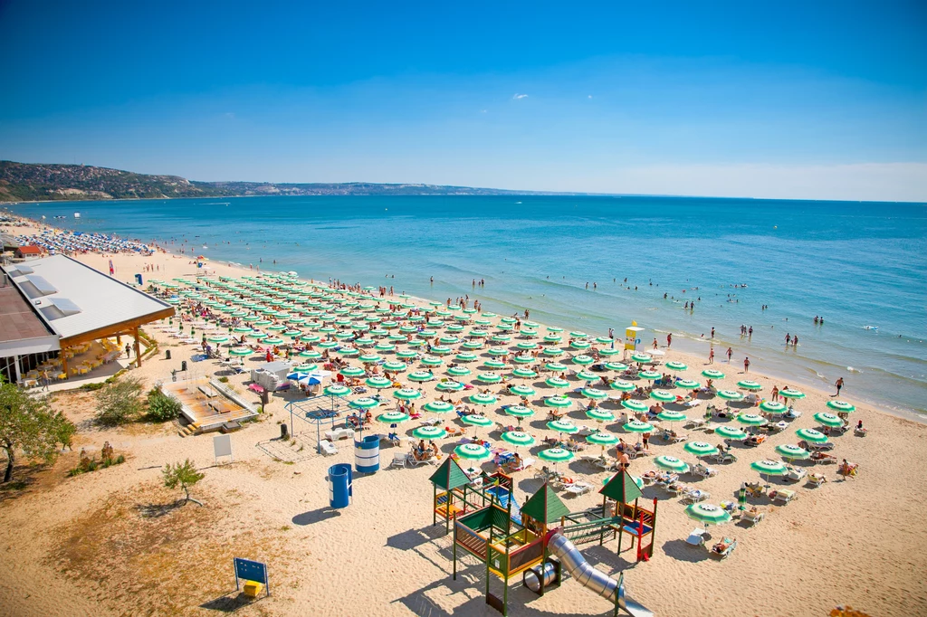 Szeroka piaszczysta plaża z licznymi parasolami i leżakami ustawionymi w rzędach, duża ilość osób korzystających z kąpieli i słonecznych dni, w tle spokojne morze oraz fragment restauracji czy baru plażowego, plac zabaw dla dzieci z kolorowymi konstruk...