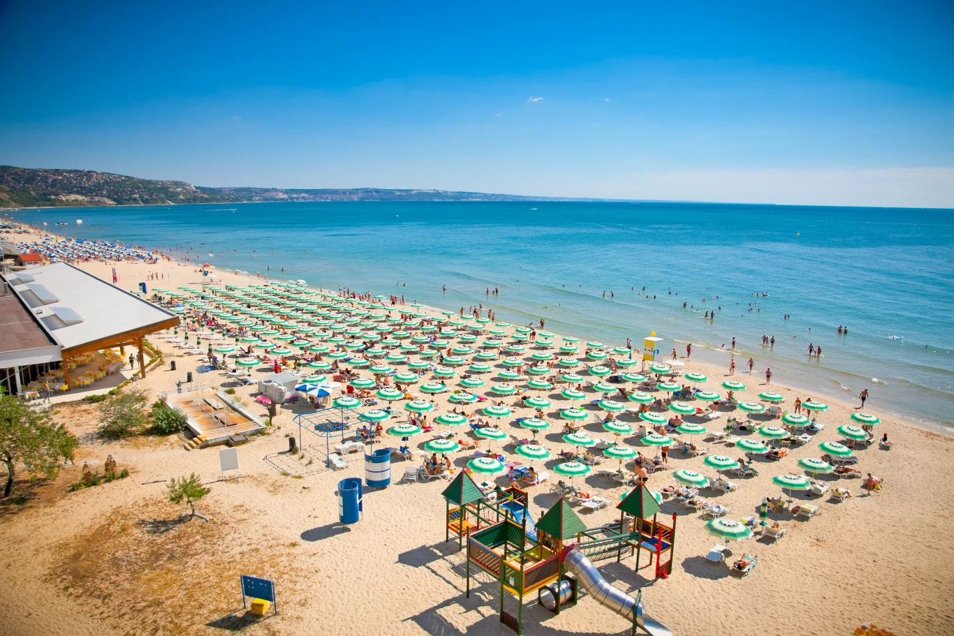W sierpniu w Bułgarii możemy wypocząć już od 800 zł Szeroka piaszczysta plaża z licznymi parasolami i leżakami ustawionymi w rzędach, duża ilość osób korzystających z kąpieli i słonecznych dni, w tle spokojne morze oraz fragment restauracji czy baru plażowego, plac zabaw dla dzieci z kolorowymi konstruk...