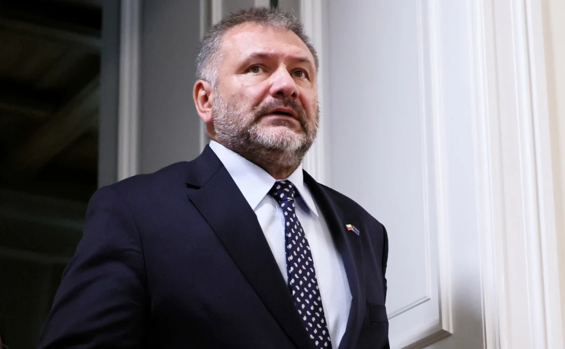 Minister sprawiedliwości Waldemar Żurek Mężczyzna w średnim wieku w ciemnym garniturze, białej koszuli i granatowym krawacie z wzorem, stoi przy białych drzwiach w jasnym wnętrzu, prezentuje poważny wyraz twarzy, ma krótko przycięte włosy i brodę.