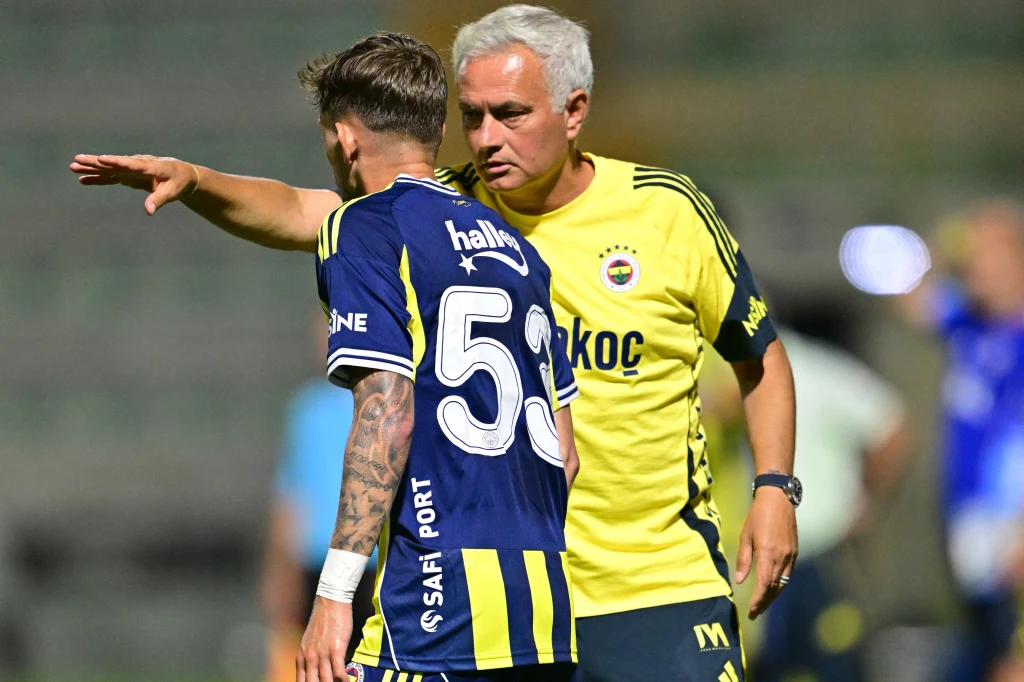 Trener w żółtej koszulce udziela wskazówek piłkarzowi w granatowym stroju z numerem 54; obaj skoncentrowani, trener wskazuje ręką kierunek na boisku, tło rozmyte, wskazuje na atmosferę meczu piłki nożnej.