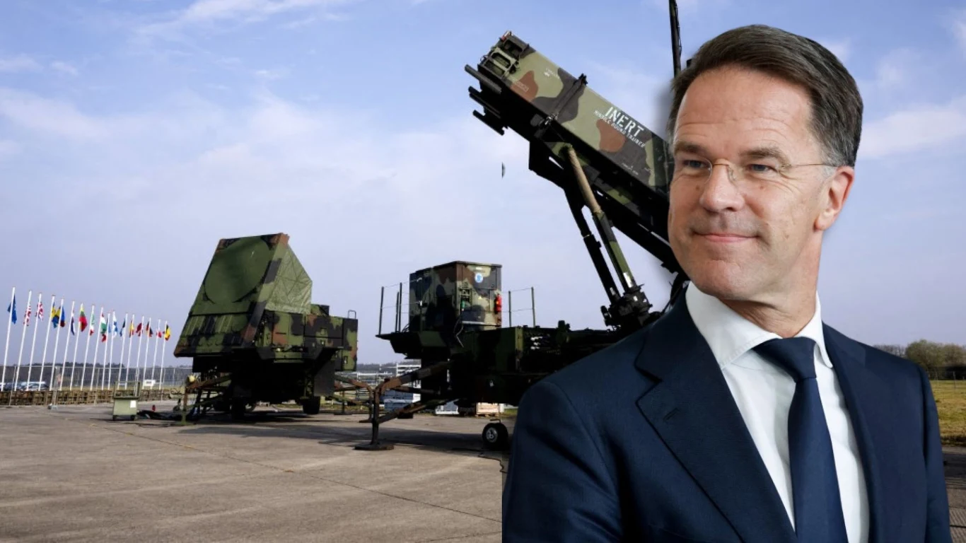 Szwecja, Dania i Norwegia dołączają do programu NATO wspierającego Ukrainę Polityk w garniturze na pierwszym planie, w tle wojskowy system rakietowy Patriot ustawiony na placu z rzędem flag państwowych po lewej stronie, pod błękitnym niebem.