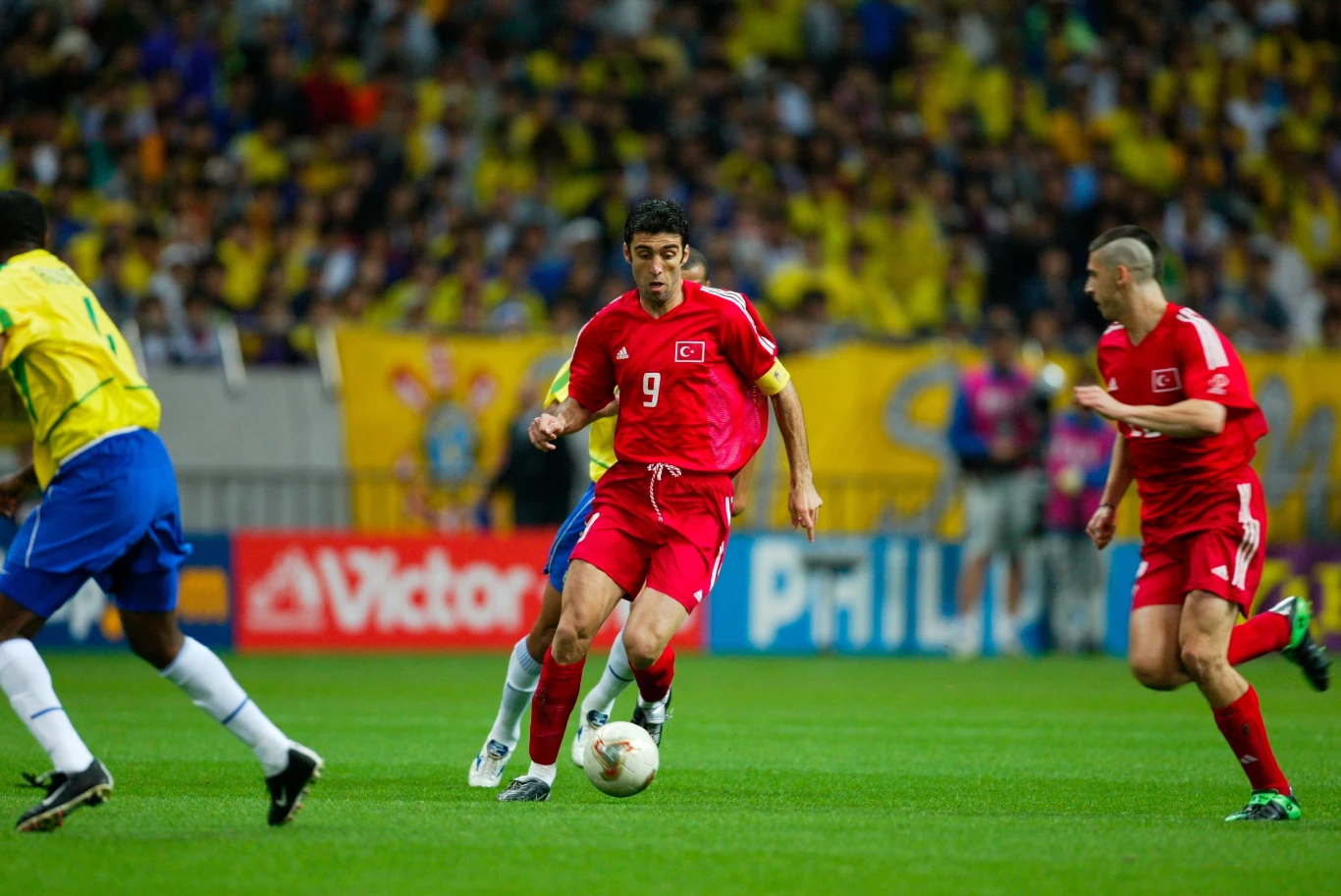 Hakan Sukur doprowadził reprezentację Turcji do brązowego medalu mistrzostw świata w 2002 roku Piłkarz w czerwonym stroju z numerem 9 prowadzi piłkę podczas meczu, a dookoła niego znajdują się inni zawodnicy na tle tłumu kibiców.