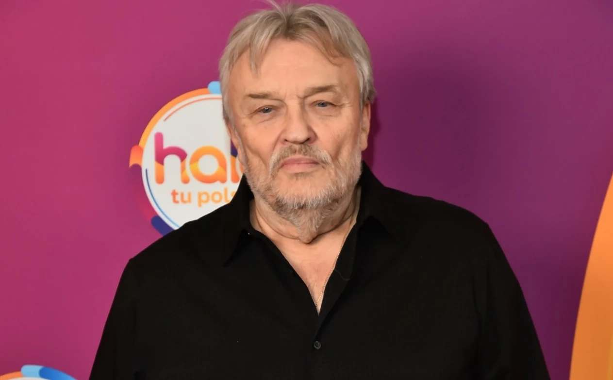 Krzysztof Cugowski odpowiedział na krytykę internautów Mężczyzna o siwych włosach i brodzie w czarnej koszuli pozuje na tle ściany z kolorowym logo i napisem.