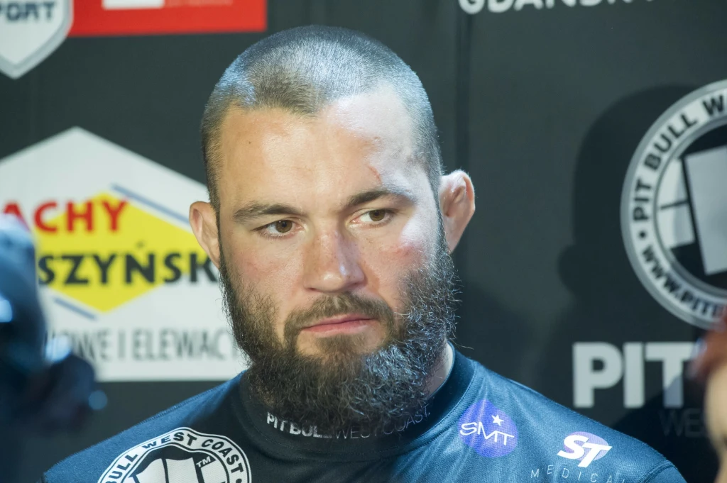 Brodaty mężczyzna w czarnej koszulce sportowej z logotypami, stojący na tle ściany z reklamami oraz logiem organizacji sportowej.